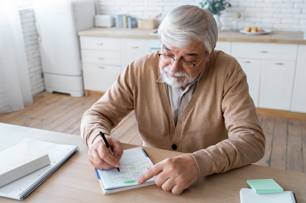 Senior man with gray hair and glasses sitting at a wooden table in a bright kitchen, writing notes in a notebook while studying or organizing his daily schedule.