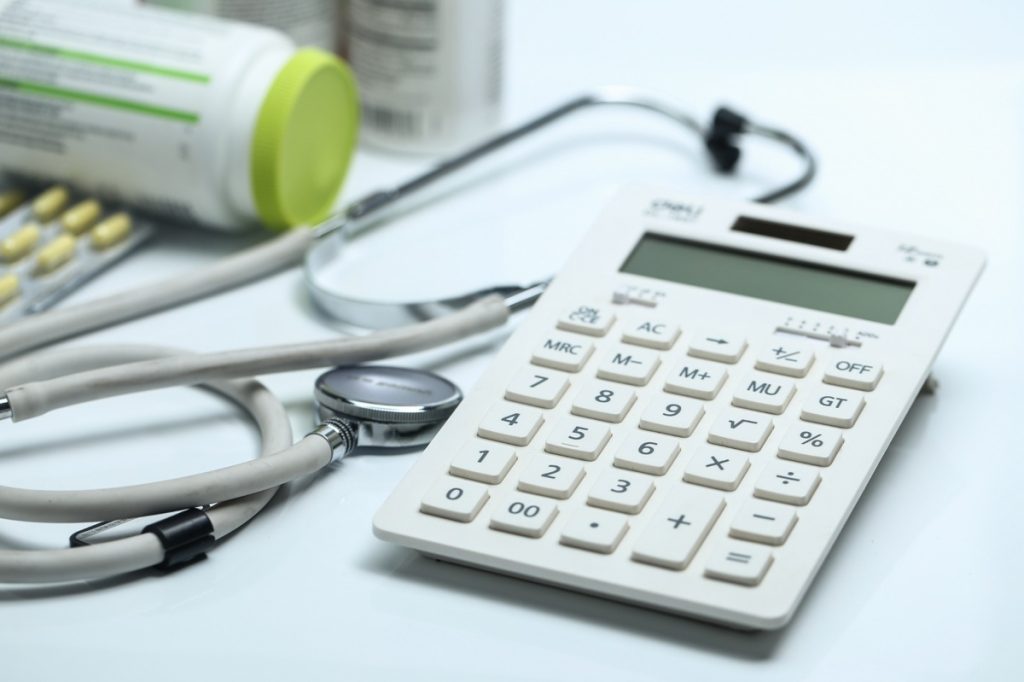 A medical-themed still life featuring a calculator, stethoscope, and pill bottles on a white surface, representing the connection between healthcare costs, budgeting, and medical expense management.