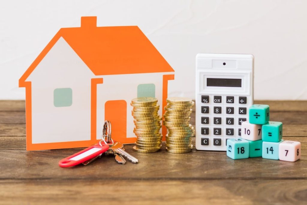 Financial composition on a wooden table showing a paper house, stacks of gold coins, house keys, a calculator, and colorful math blocks, symbolizing budgeting, housing costs, and financial planning related to rates and home expenses.