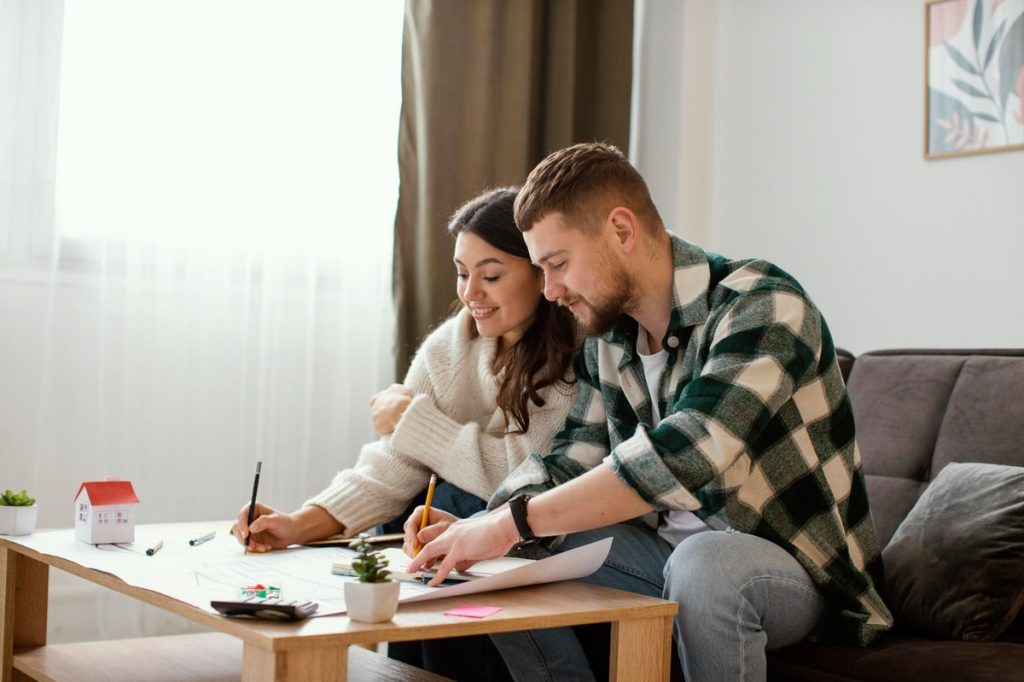 Young couple sitting together on a couch at home, smiling while writing and reviewing papers on a wooden coffee table decorated with small plants and a miniature house model.