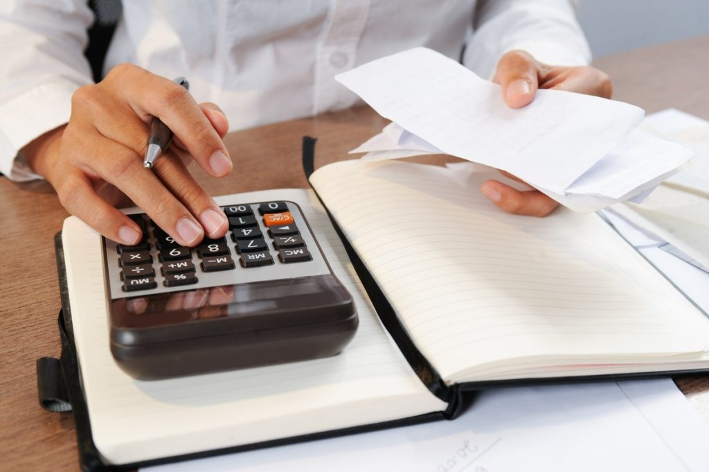 Person holding receipts and pressing buttons on a calculator while writing notes in a notebook, managing personal or business finances with focus and concentration.