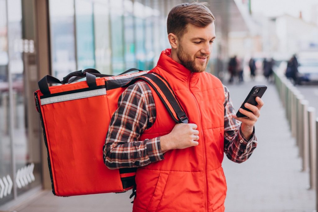 A man wearing a red delivery vest and plaid shirt carries an insulated food delivery bag on his back while checking his smartphone on a city sidewalk, representing the gig economy workforce in action.
