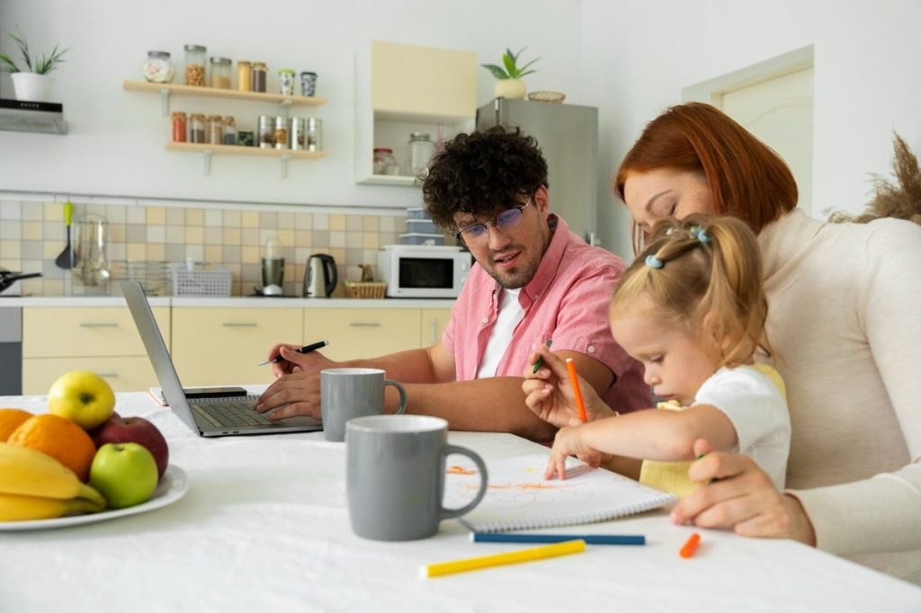 A young couple sits at a kitchen table with their daughter, engaging in different activities together. The father works on a laptop while taking notes, the mother helps the little girl draw with colored pencils, and a bowl of fresh fruit and coffee mugs sit on the table. The scene conveys a warm, balanced home environment where family and financial responsibilities are managed together.